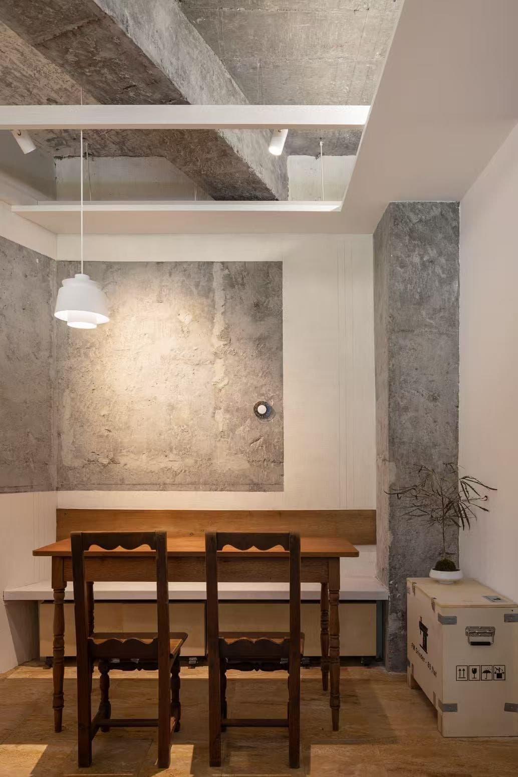 A dark long table, two wooden chairs and a white pendant in front of a concrete wall, with a column and a plant beside